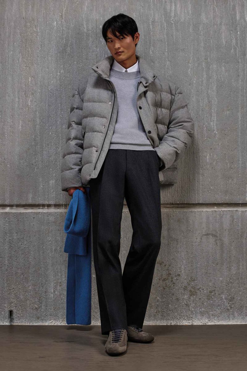 Model wearing a grey puffer jacket and dark trousers standing against a textured concrete wall