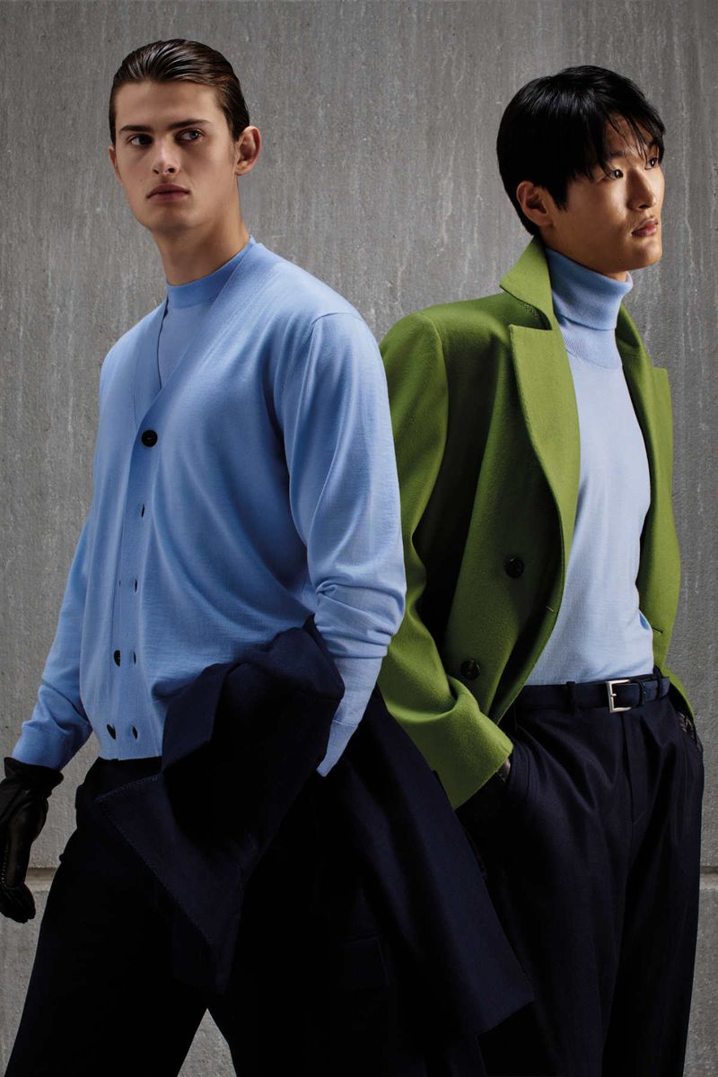 Two male models posing against a textured grey wall for Kiton, wearing knitwear and tailored outerwear.