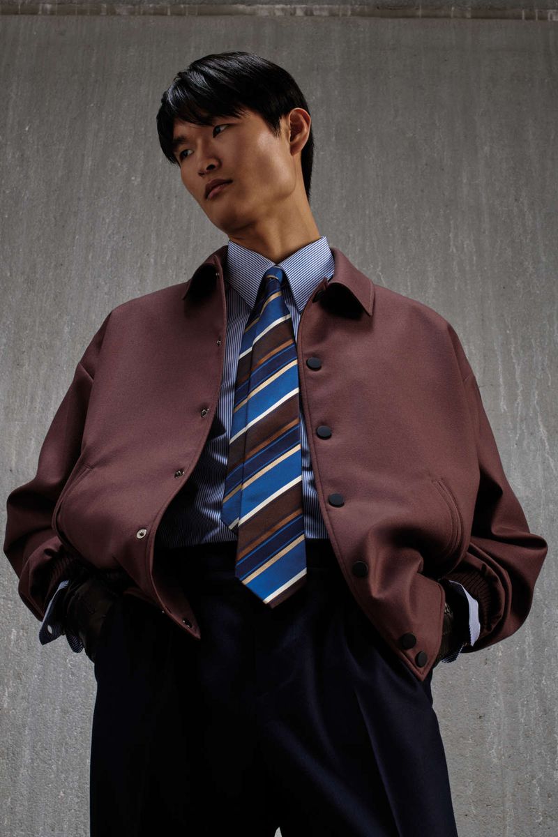 Asian male model wearing a maroon jacket, striped shirt, and diagonal striped tie against a concrete wall