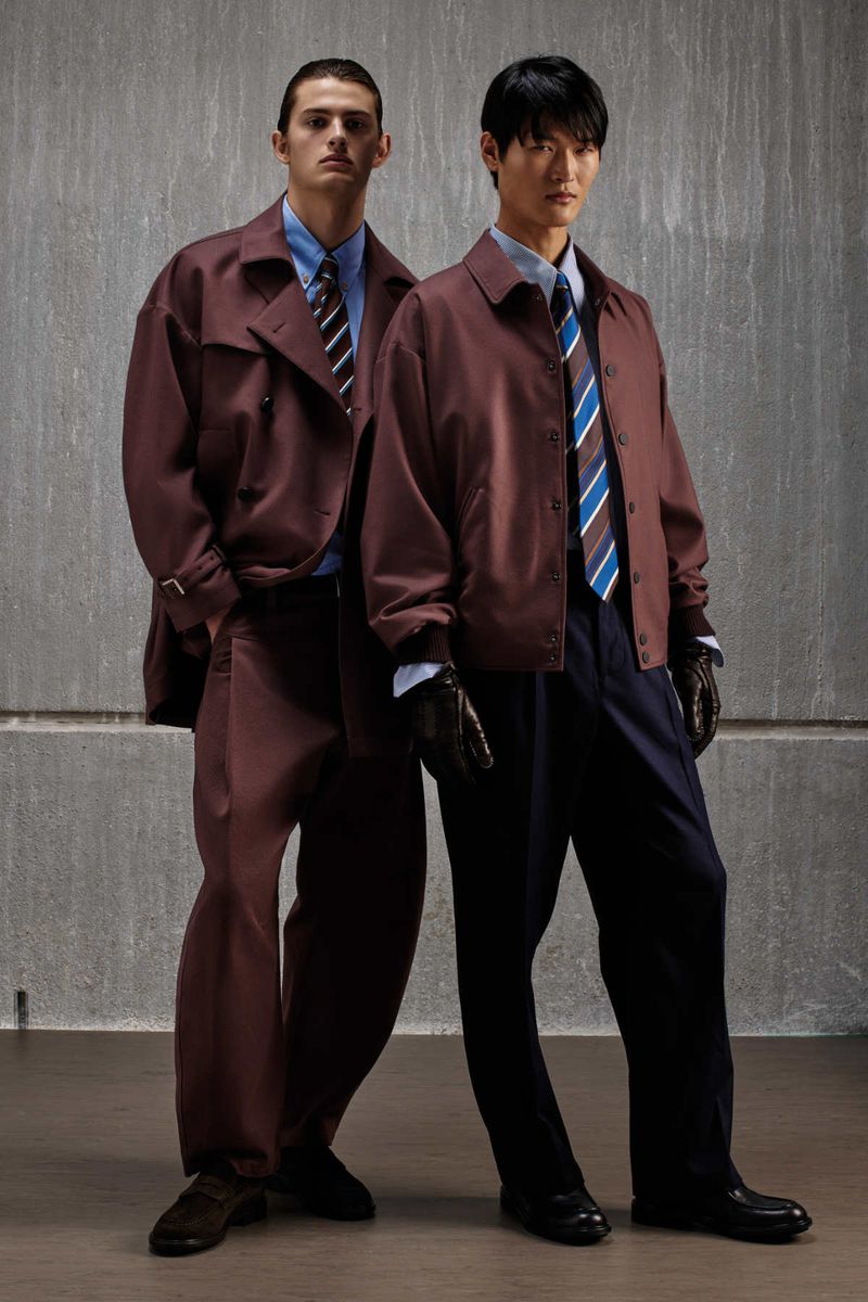 Two male models posing against a textured concrete wall wearing matching maroon jackets, blue shirts, and striped ties.