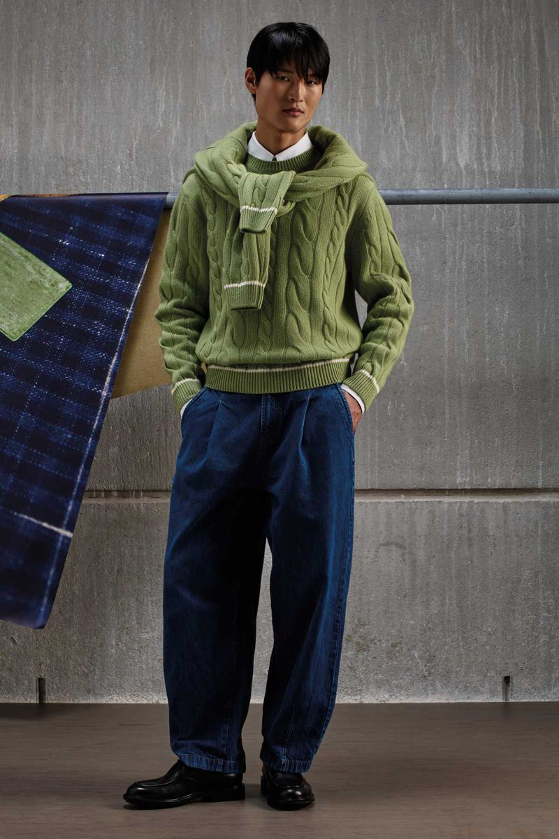 Asian man wearing a green cable-knit sweater, blue denim trousers, and dark shoes against a concrete wall.