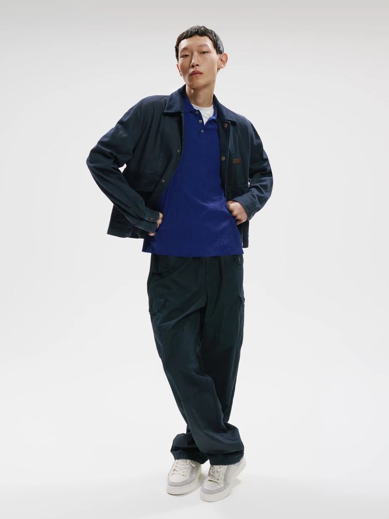 Model wearing a dark jacket, blue polo shirt, and dark trousers against a white seamless background
