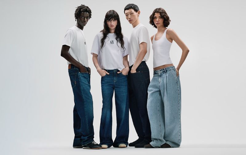Four models standing against a plain white background wearing white t-shirts and blue denim jeans for Armani Exchange.