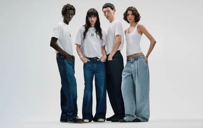 Four models standing against a plain white background wearing white t-shirts and blue denim jeans for Armani Exchange.
