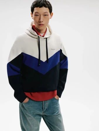 Model wearing a color-blocked hoodie in white, blue, and black over a red shirt with blue jeans against a white background