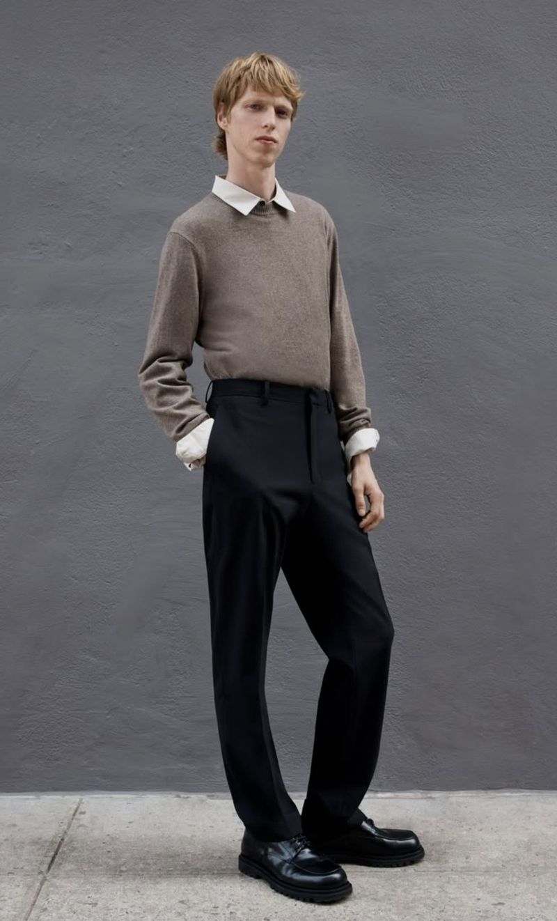 A male model wearing a brown knit sweater over a white collared shirt and black trousers against a textured grey wall.