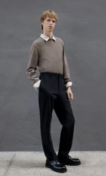 A male model wearing a brown knit sweater over a white collared shirt and black trousers against a textured grey wall.