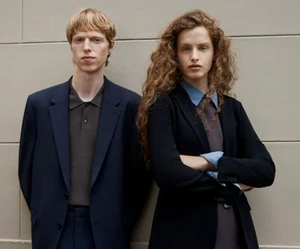 Two models standing side by side wearing tailored jackets against a plain stone wall background