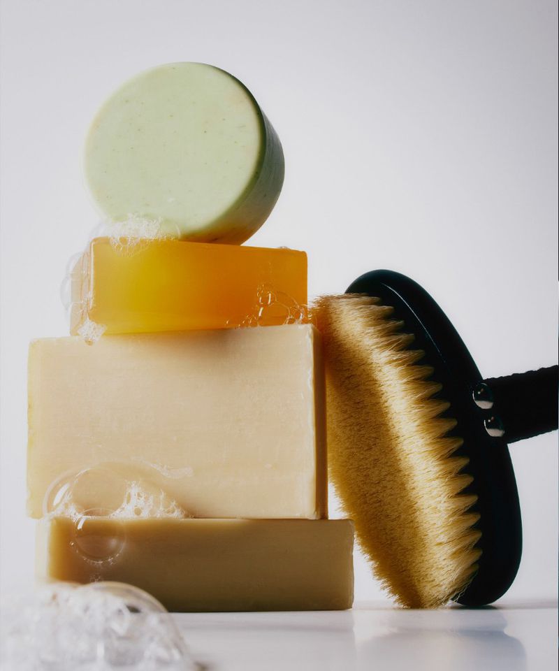 Stacked colorful soaps with visible lather bubbles and a black wooden body brush against a plain white background