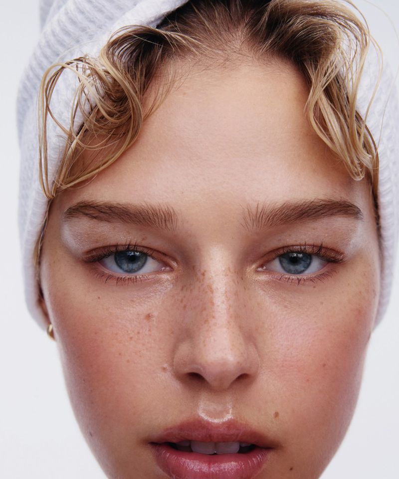 Close-up portrait of a young woman with freckles wearing a white towel on her head against a seamless white background.