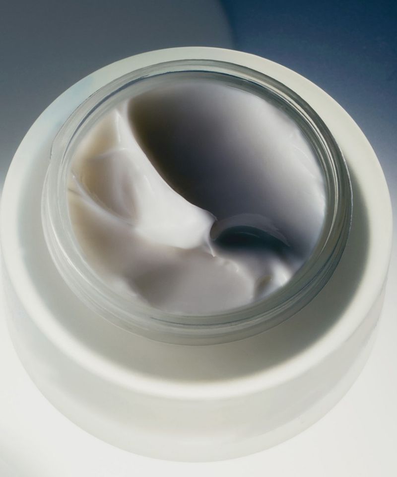Top-down view of an open jar of skincare cream with smooth white and grey swirled textures set on a bright white surface.