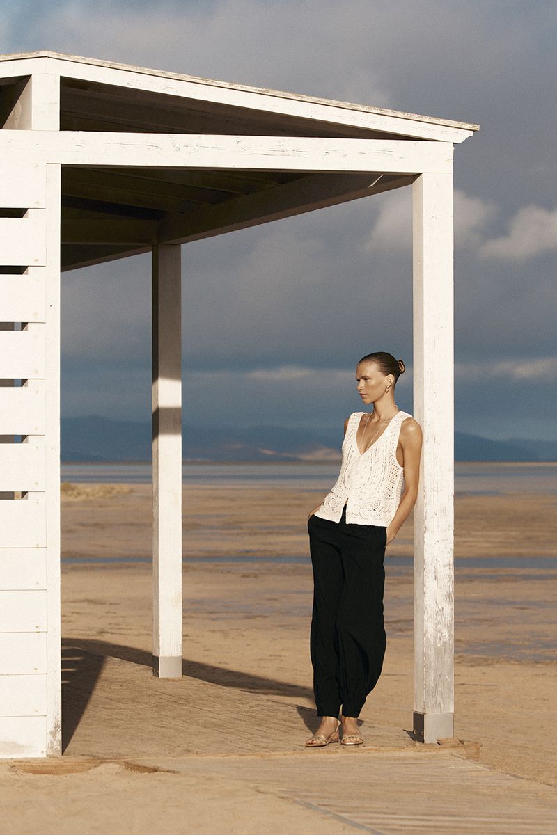 Model leaning against a white wooden structure on a sandy beach wearing a knit top and dark trousers.