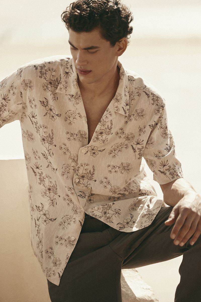 Male model wearing a patterned floral button-down shirt and dark trousers against a neutral background.