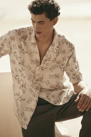 Male model wearing a patterned floral button-down shirt and dark trousers against a neutral background.