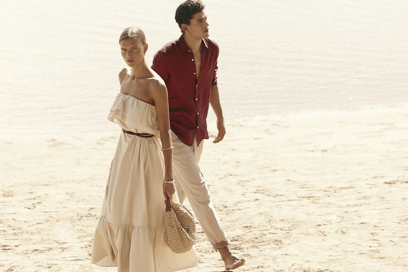 A woman in a cream strapless dress and a man in a red shirt walking together on a sandy beach.
