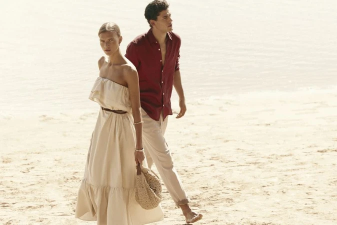 A woman in a cream strapless dress and a man in a red shirt walking together on a sandy beach.