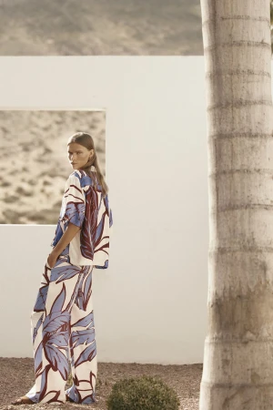 Model standing outdoors near a white wall wearing a blue and white patterned shirt and matching trousers.