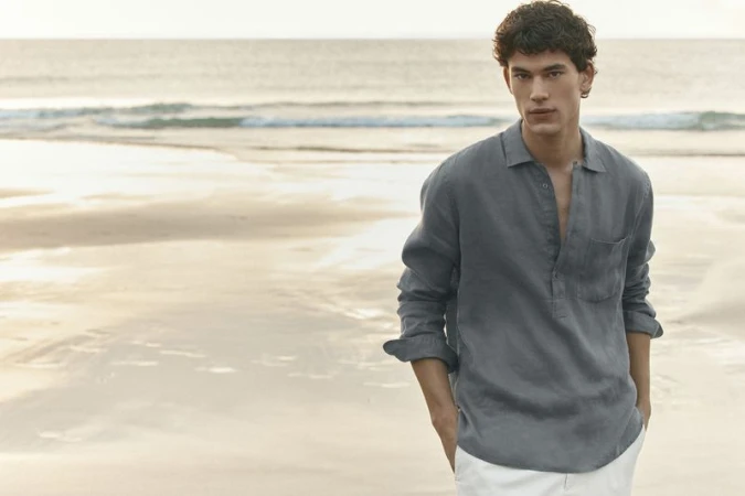Male model wearing a grey linen shirt standing on a sandy beach by the ocean for Pedro del Hierro.