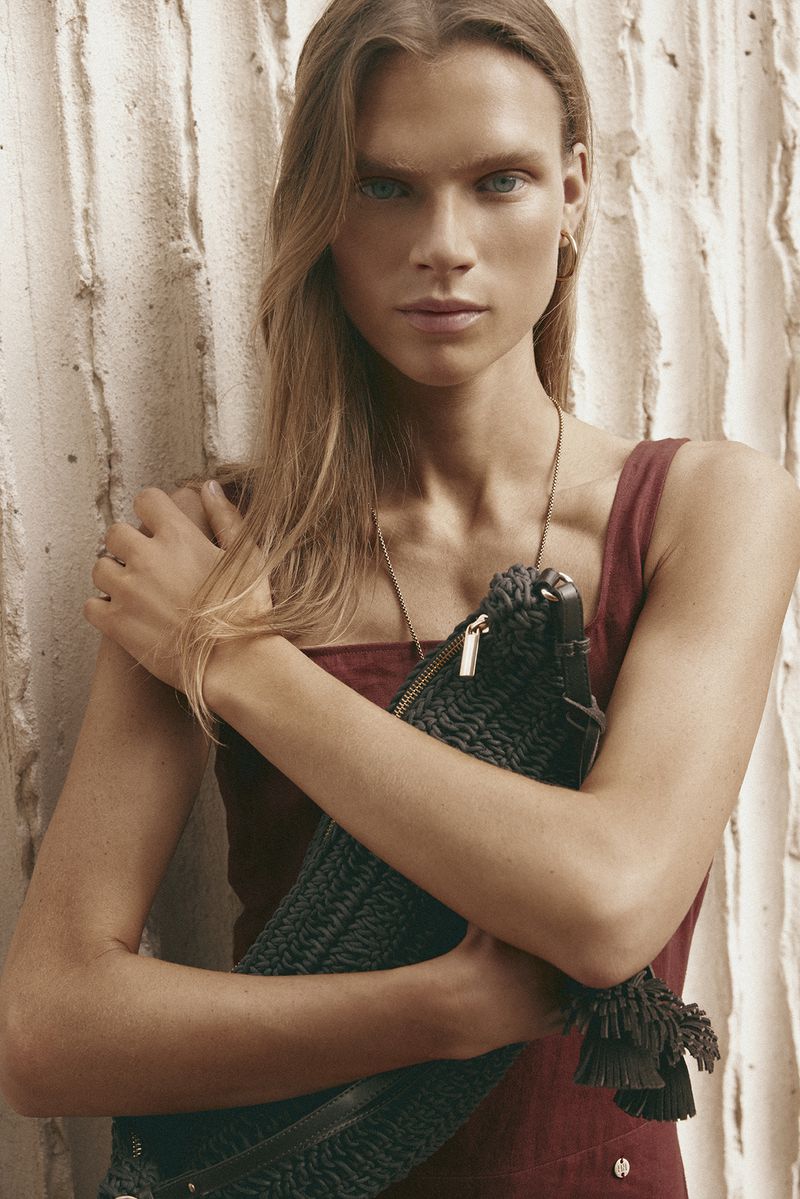 Model with long blonde hair holding a dark woven handbag wearing a burgundy top against a textured white wall