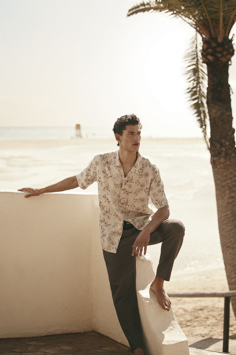 A male model wearing a patterned short-sleeve shirt and dark trousers leaning against a low wall near a beach and palm tree.