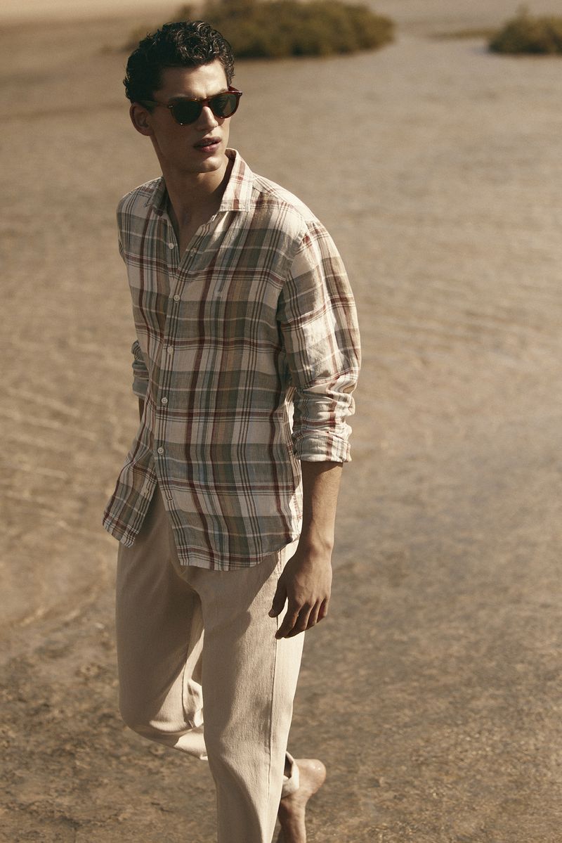 Male model in a plaid shirt and sunglasses walking on a sandy beach for Pedro del Hierro.