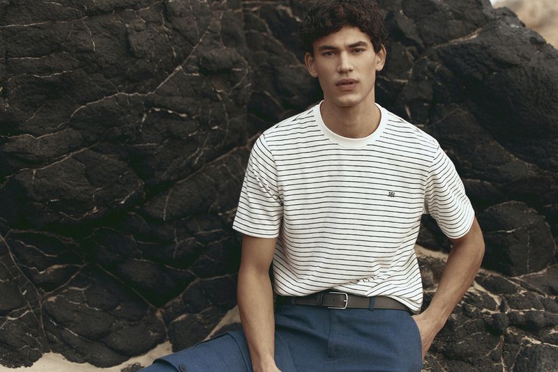Male model wearing a striped t-shirt and blue trousers seated against dark rocky coastal cliffs.