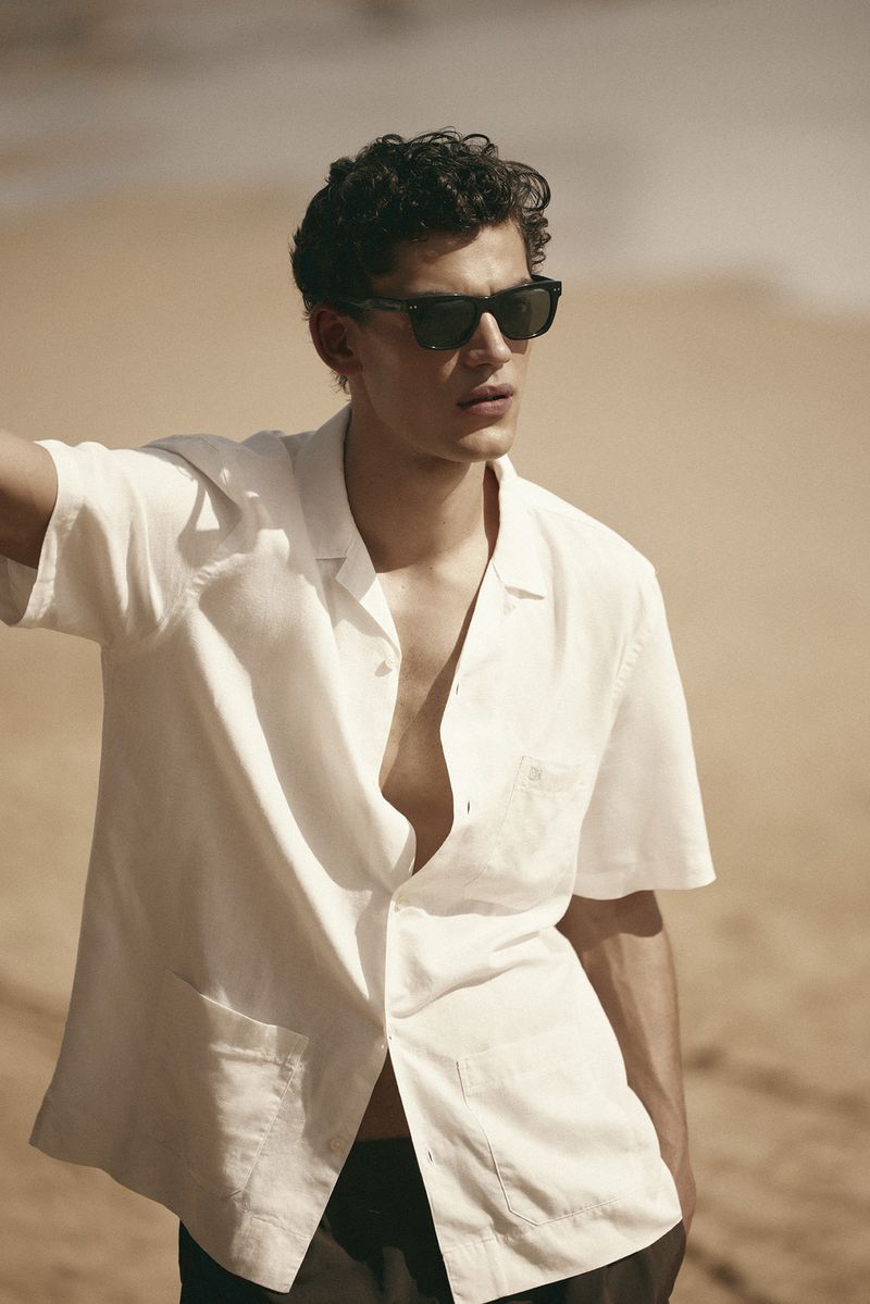 A male model wearing black sunglasses and a white short-sleeve button-down shirt standing against a sandy beach background.