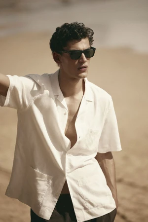 A male model wearing black sunglasses and a white short-sleeve button-down shirt standing against a sandy beach background.