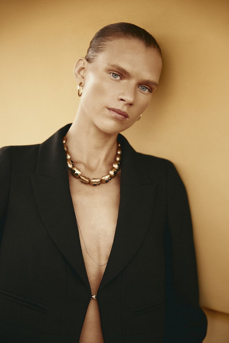 Model in a black jacket wearing a gold statement necklace against a warm yellow backdrop