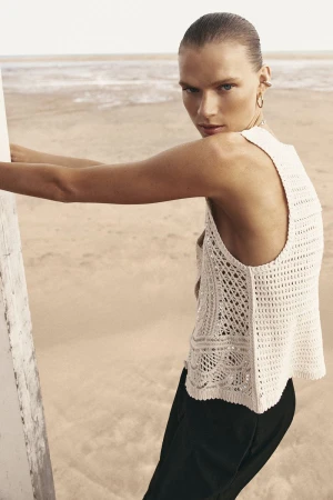 Model wearing a white knit top and dark pants leaning against a structure with a sandy beach background.