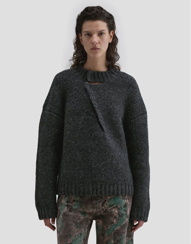 Model wearing a dark charcoal wool sweater with an asymmetric neckline against a plain light grey studio backdrop.