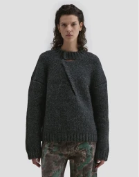 Model wearing a dark charcoal wool sweater with an asymmetric neckline against a plain light grey studio backdrop.