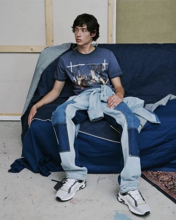 Young male model seated on a dark blue sofa wearing a printed graphic t-shirt and patchwork denim jeans for Off White.