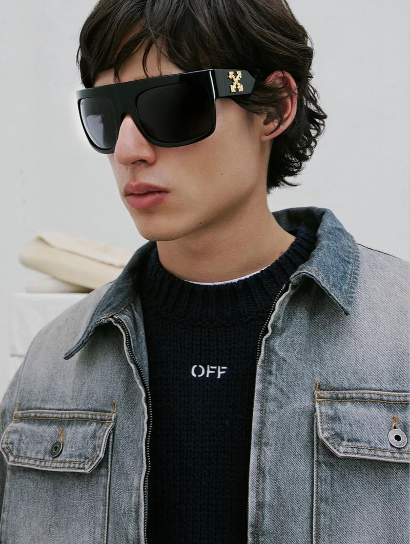 Model wearing black sunglasses with gold branding and a denim jacket over a dark sweater against a white wall.