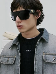 Model wearing black sunglasses with gold branding and a denim jacket over a dark sweater against a white wall.