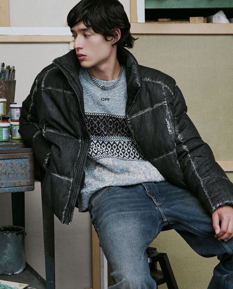 Model wearing a black puffy jacket and patterned sweater seated in an artist studio with paint jars.