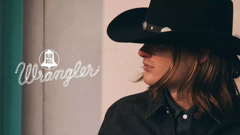 Close-up profile of a person wearing a black cowboy hat and dark denim jacket with Wrangler Blue Bell campaign text visible on the left.