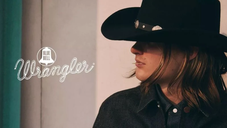 Close-up profile of a person wearing a black cowboy hat and dark denim jacket with Wrangler Blue Bell campaign text visible on the left.