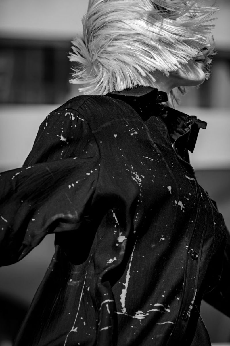 Monochromatic shot of a fair-skinned adult with short blonde hair and a black-and-white splatter-patterned shirt in motion outdoors.
