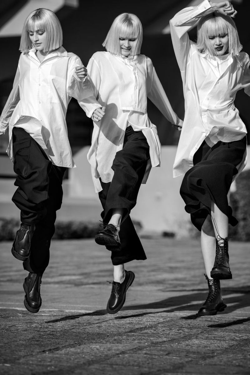 Three women with blonde bobs are holding hands and leaping in the air wearing oversized white shirts, dark pants, and black dress shoes.