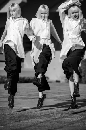 Three women with blonde bobs are holding hands and leaping in the air wearing oversized white shirts, dark pants, and black dress shoes.