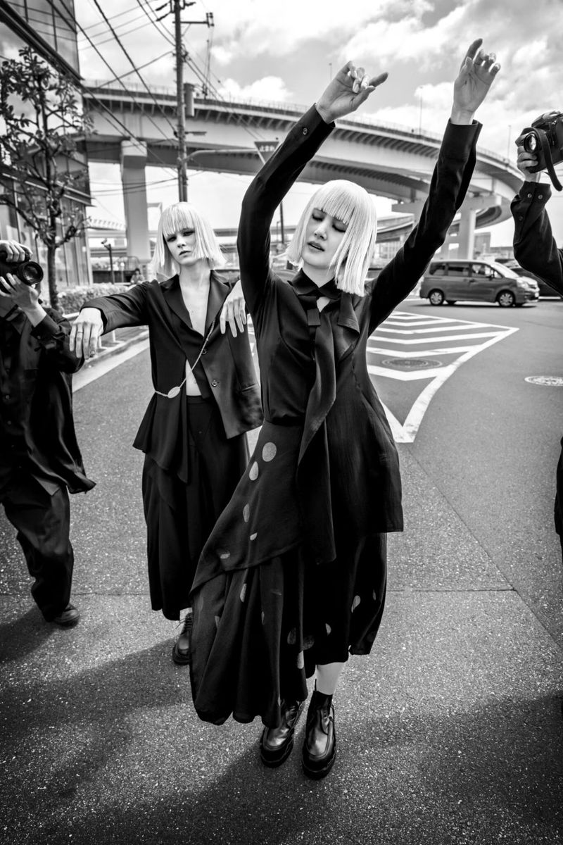 A monochrome fashion shot of two models wearing dark clothing, standing on a city street under an elevated highway with a car passing by.
