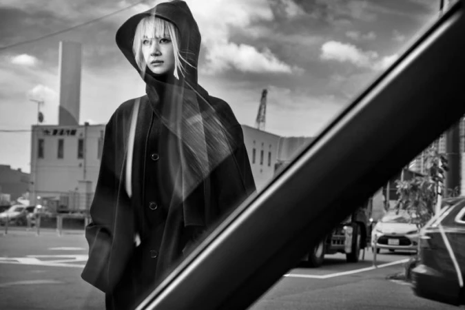 Monochrome outdoor portrait of a blonde woman in a hooded coat, captured from a car's perspective with city buildings in the background.