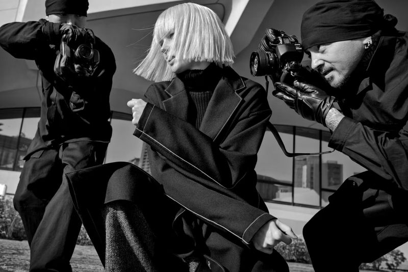 Black and white fashion shot featuring a model in a dark coat, posed in front of two photographers with cameras, in an outdoor setting.
