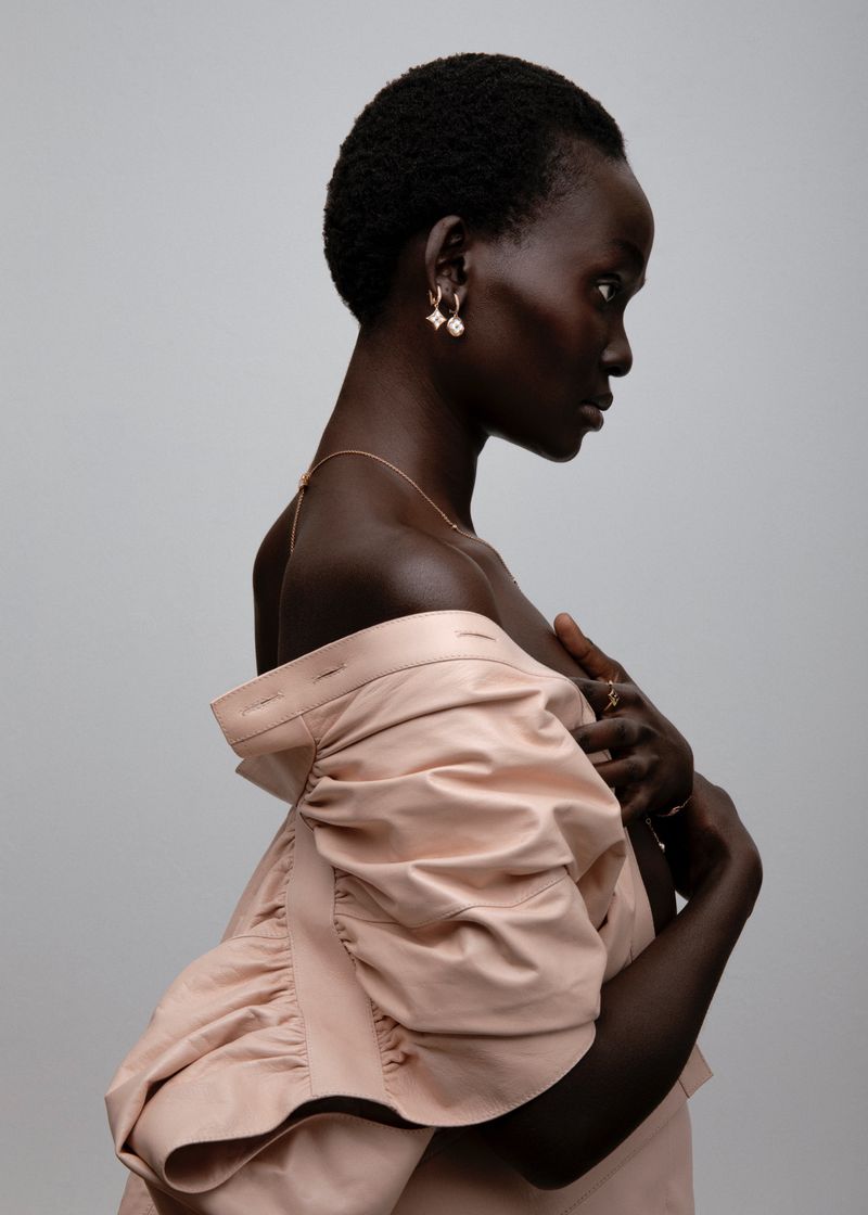 Model in profile wearing an off-the-shoulder pale pink garment against a neutral grey studio background