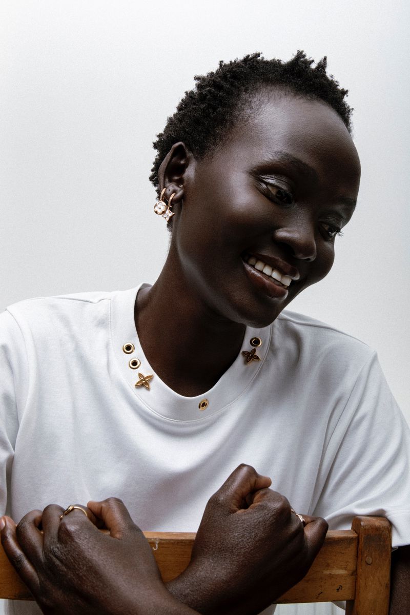 Close-up portrait of a Black woman wearing a white t-shirt with gold hardware details and earrings against a plain white backdrop