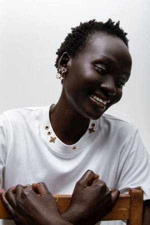Close-up portrait of a Black woman wearing a white t-shirt with gold hardware details and earrings against a plain white backdrop