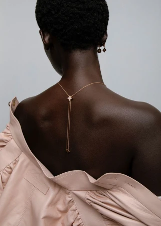 Model seen from behind wearing a pale pink jacket and delicate gold pendant necklace against a light grey background.
