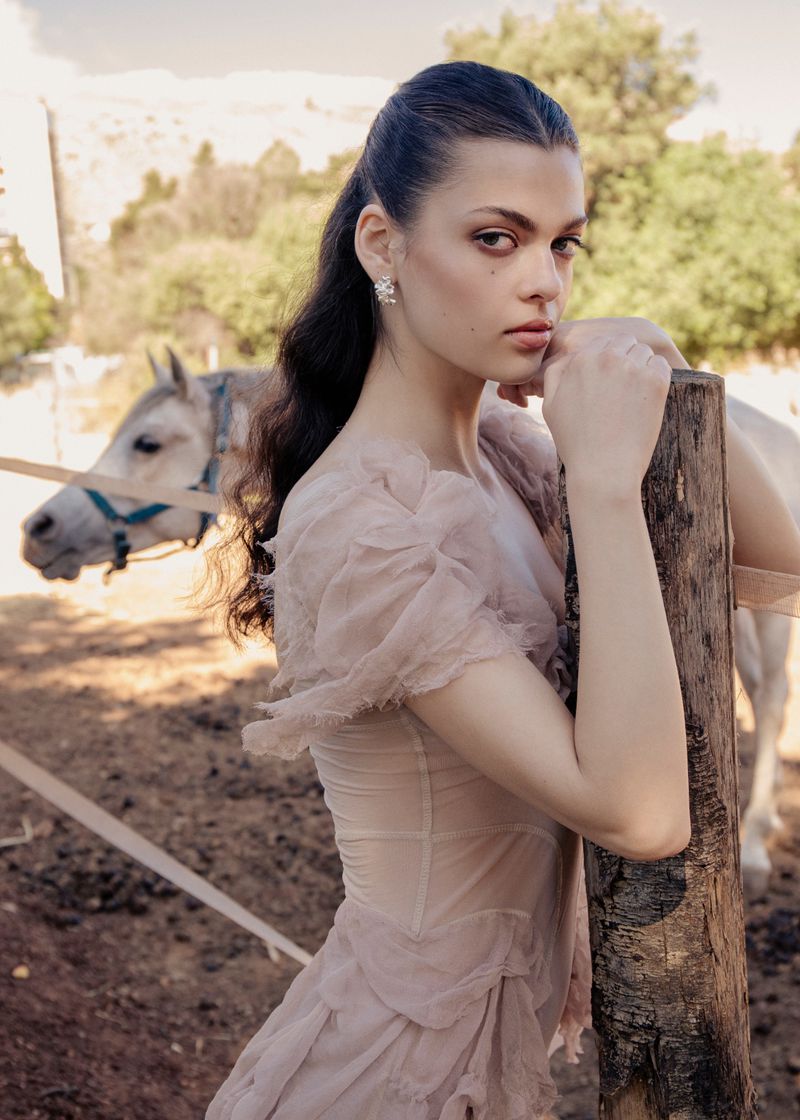 Model in pale pink dress leaning against a wooden post outdoors with a white horse standing in the background.