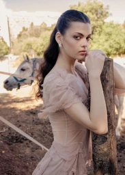 Model in pale pink dress leaning against a wooden post outdoors with a white horse standing in the background.